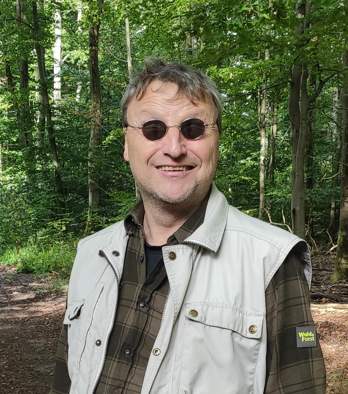 Per Busch, stehend im Wald. Er lächelt und trägt eine verspiegelte Sonnenbrille mit kleinen, kreisrunden Gläsern. (2023)